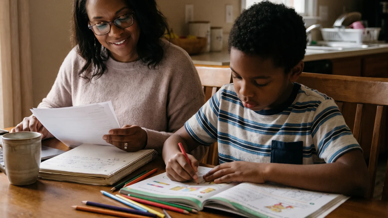 Mother reviewing a household budget with a laptop and notebook at a kitchen table while her young son does homework beside her